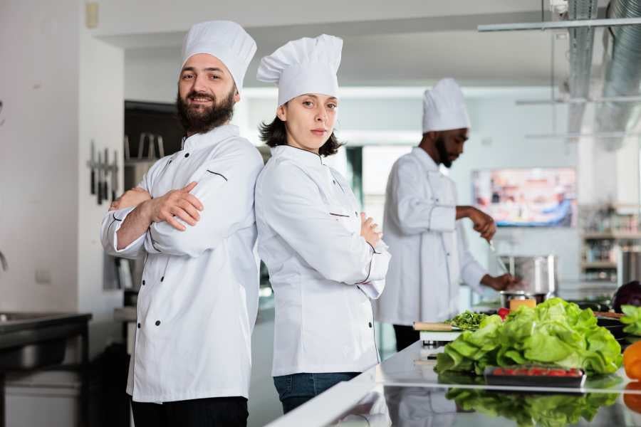 Two chefs back to back pose with a busy chef behind.