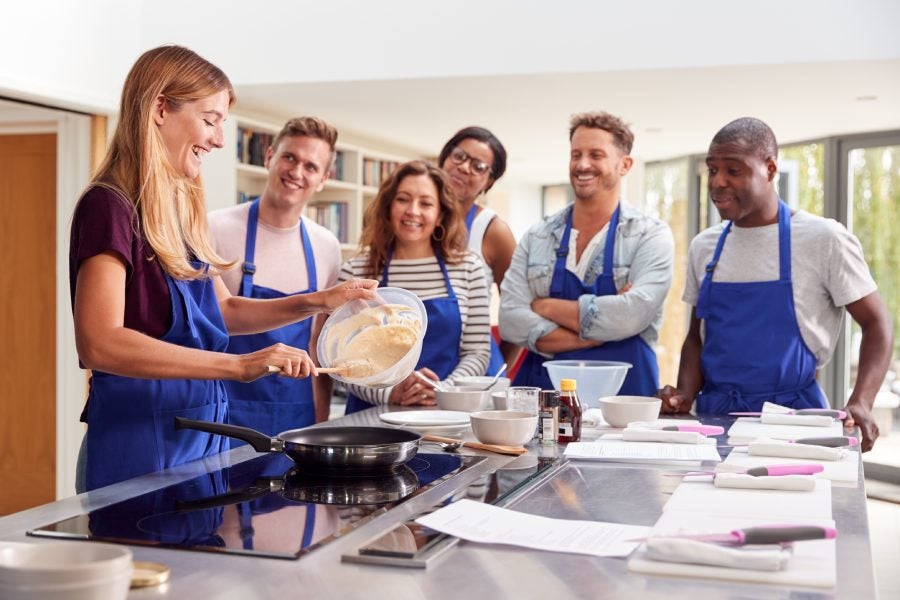 Female teacher making pancake on cooker in cookery class as adult students look on.