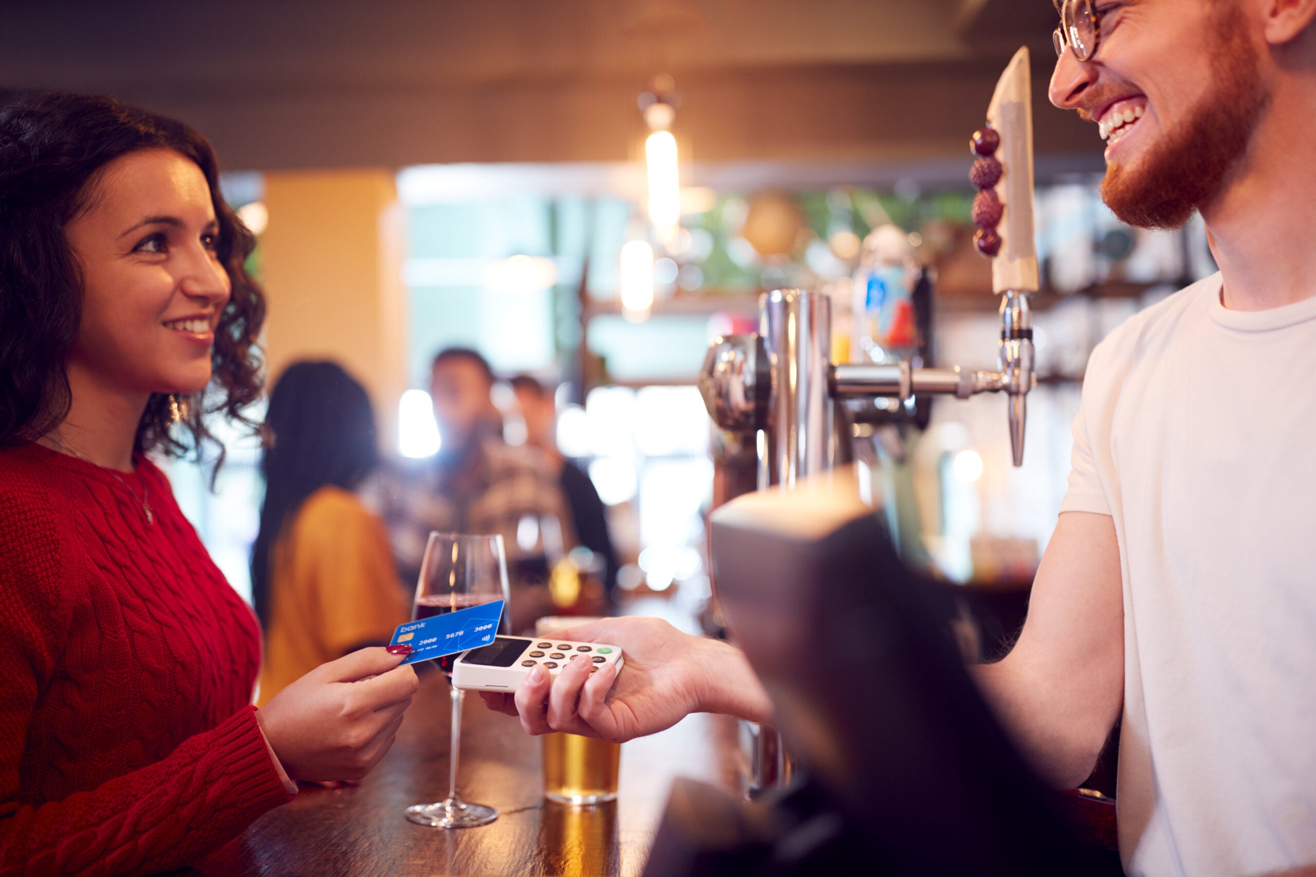 Smiling Female Customer In Bar Making Contactless Payment With Card For Drinks To Male Bartender